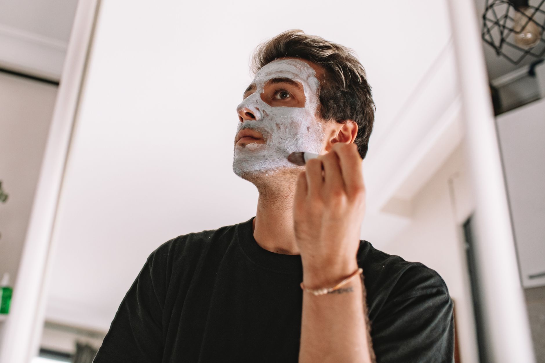 Adult man applying a facial mask while looking in the mirror at home, promoting skincare routine.