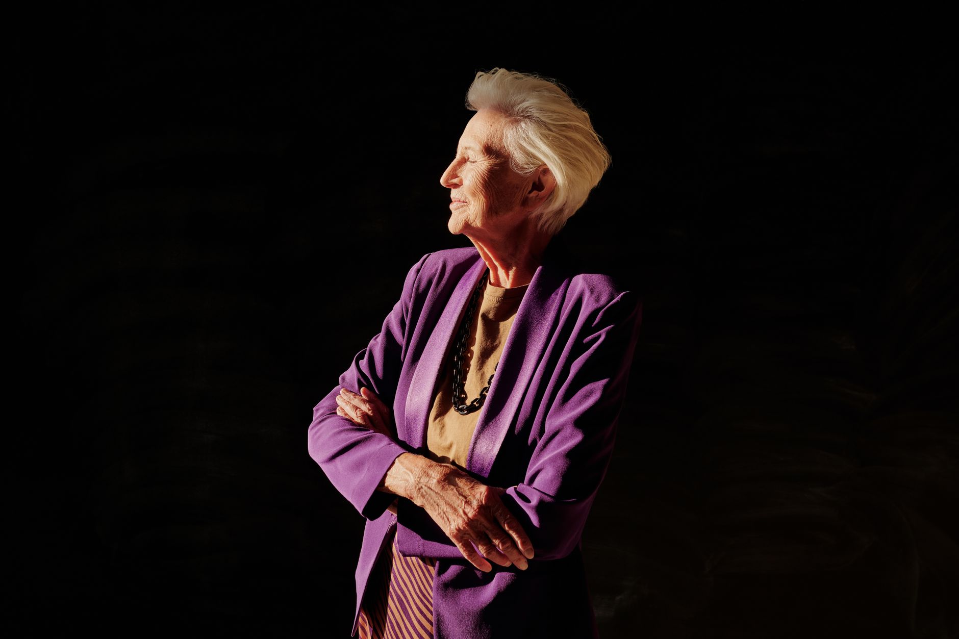 Portrait of an elderly woman in a purple blazer with arms crossed, set against a dark background.