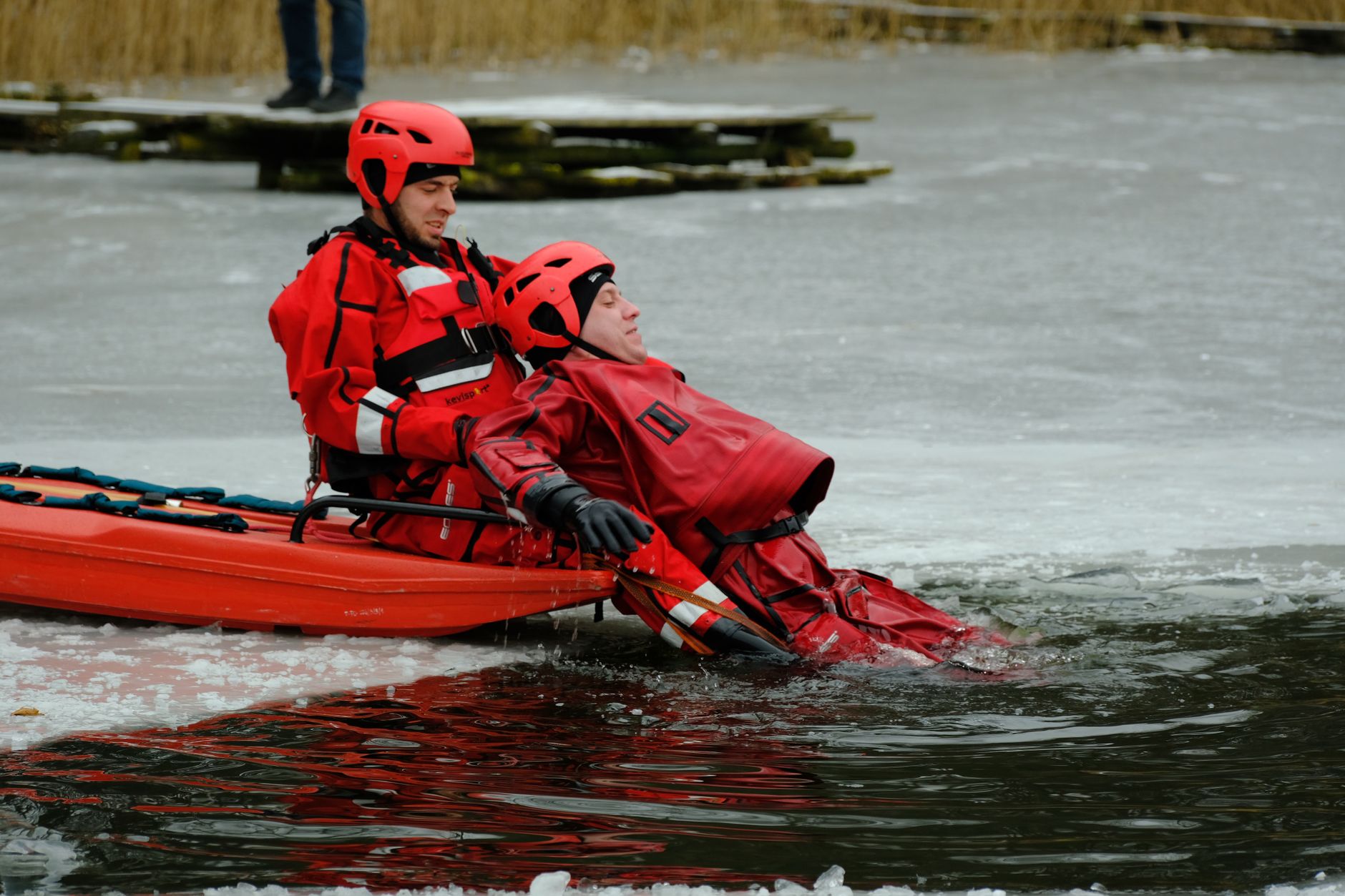 Two firefighters in red suits conduct an ice rescue drill on a frozen lake.