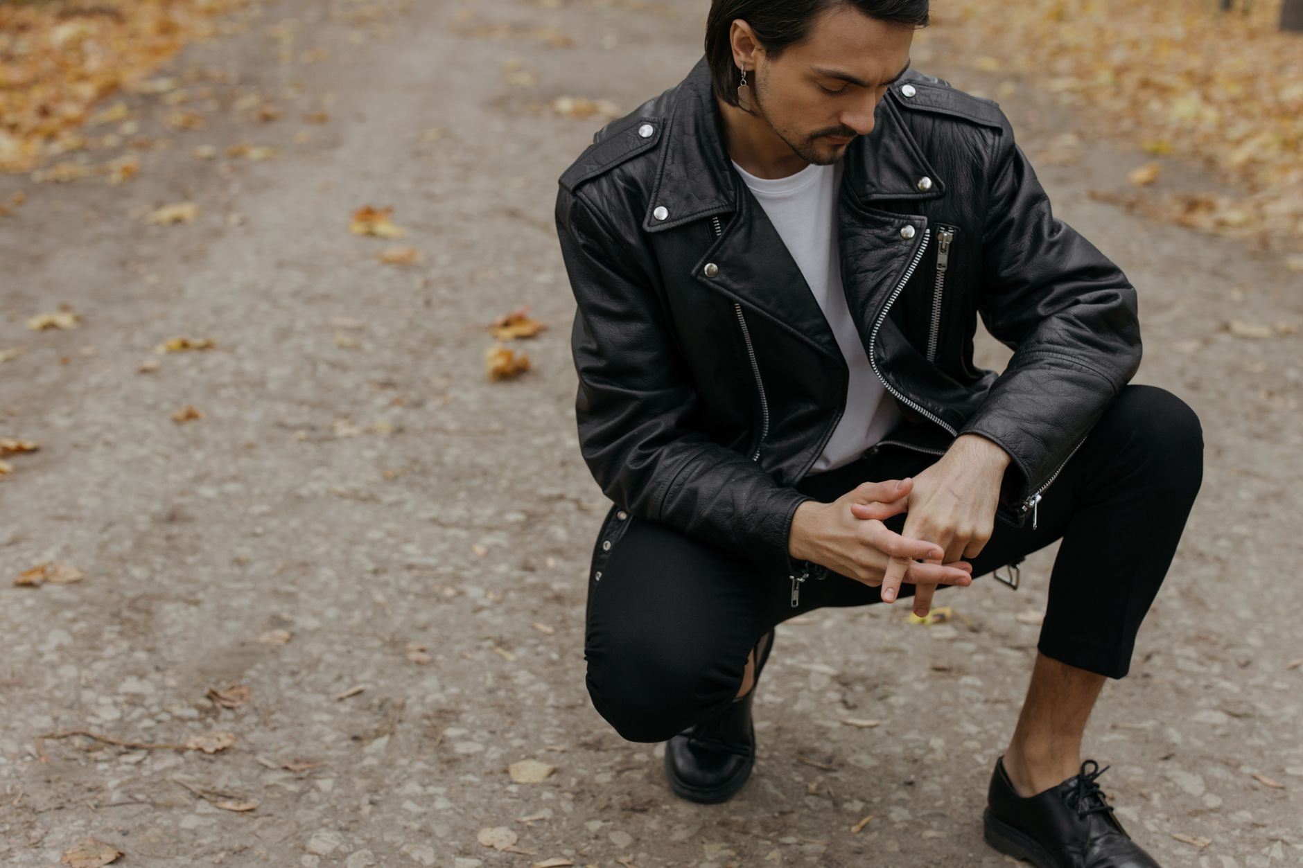 Man squatting on a path wearing a leather jacket, capturing autumn fashion style.