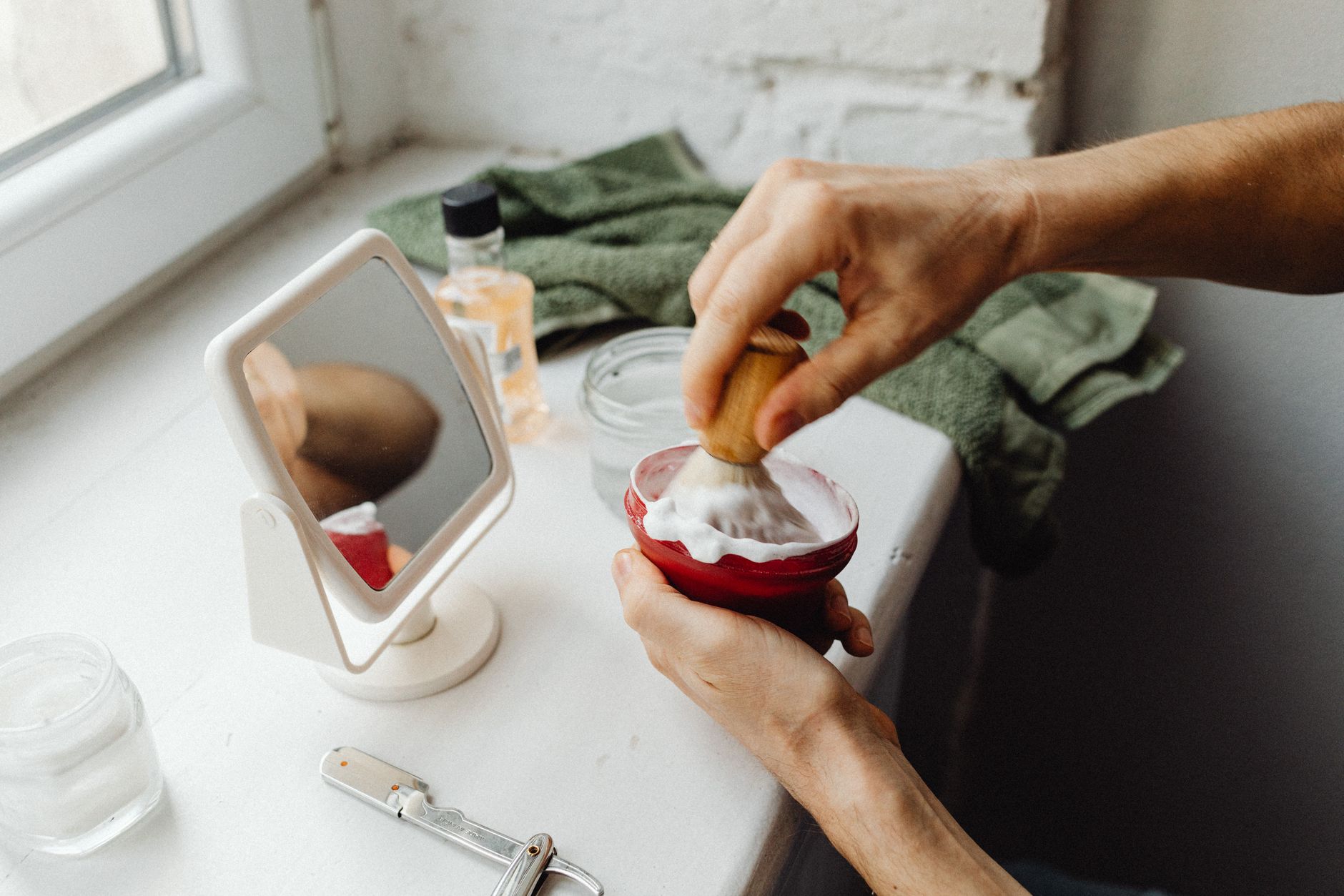 A close-up image of hands preparing shaving cream with a brush in a bathroom setting.