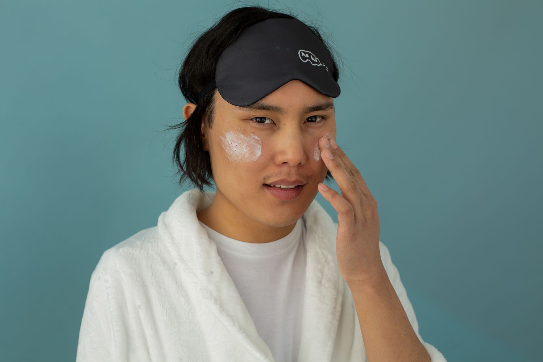 Asian man in a robe applying skincare face cream for facial care with a sleep mask.