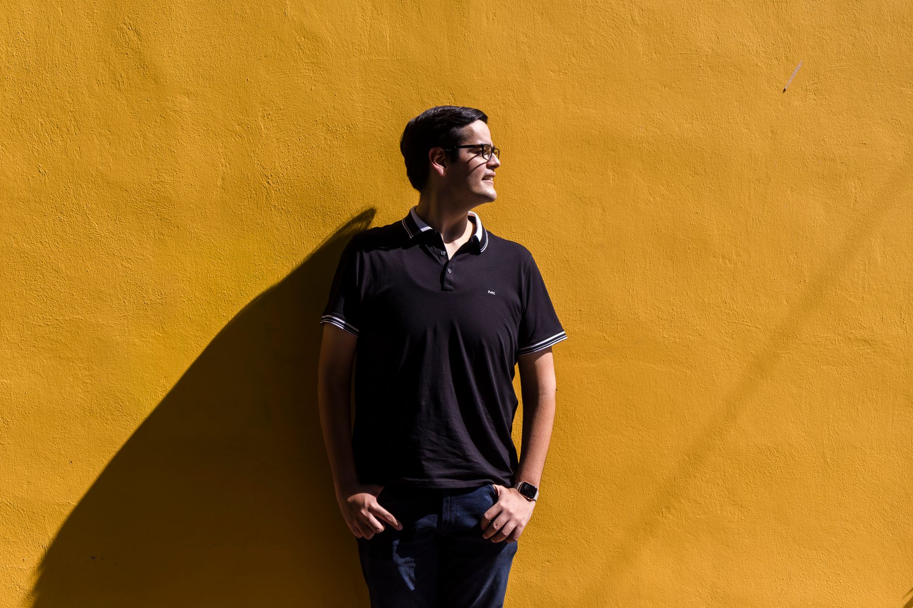 A man in casual black attire poses confidently against a vibrant yellow wall, casting a striking shadow.