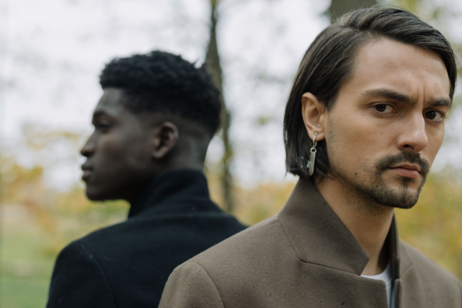 Two men in stylish autumn fashion, one facing forward, one turned away, outdoors.