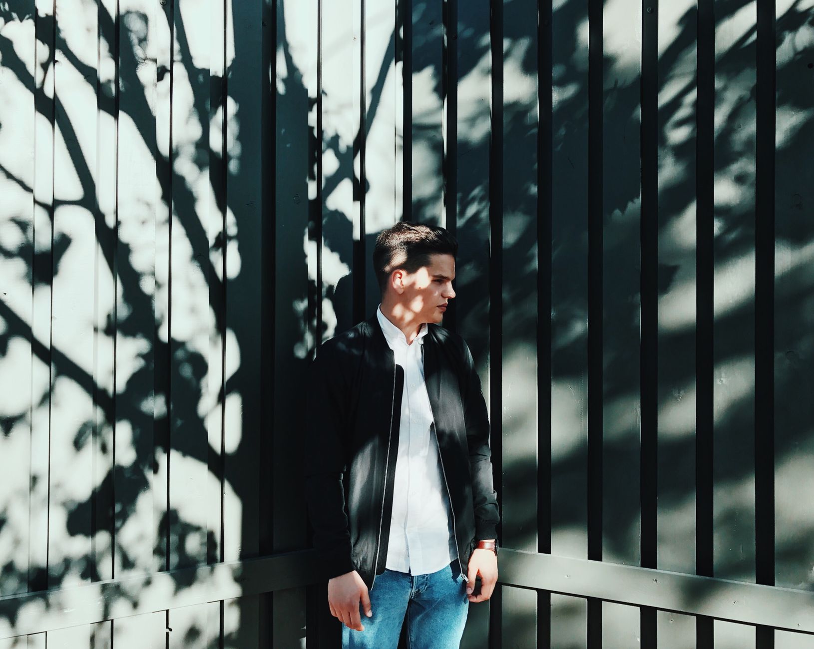 A young man in a black bomber jacket stands outdoors, with dramatic shadows.