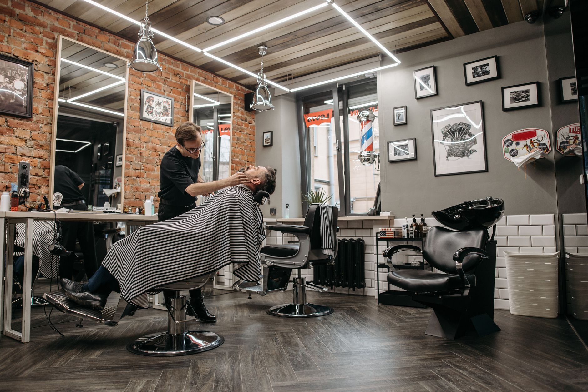 A barber attends to a client in a stylish, contemporary barbershop with brick walls and framed art.