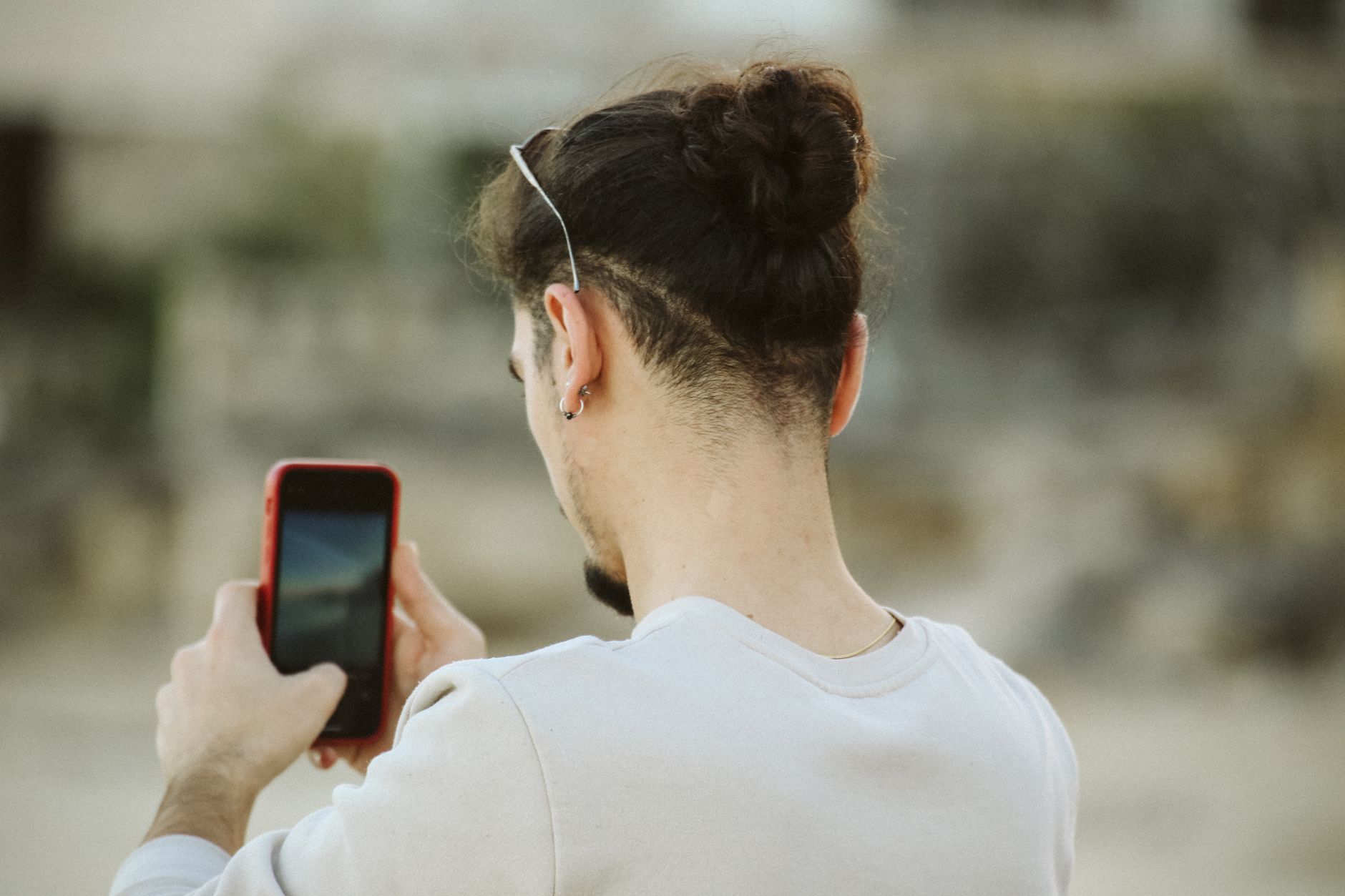 A young man with a bun hairstyle captures a photo using his smartphone in an outdoor setting.