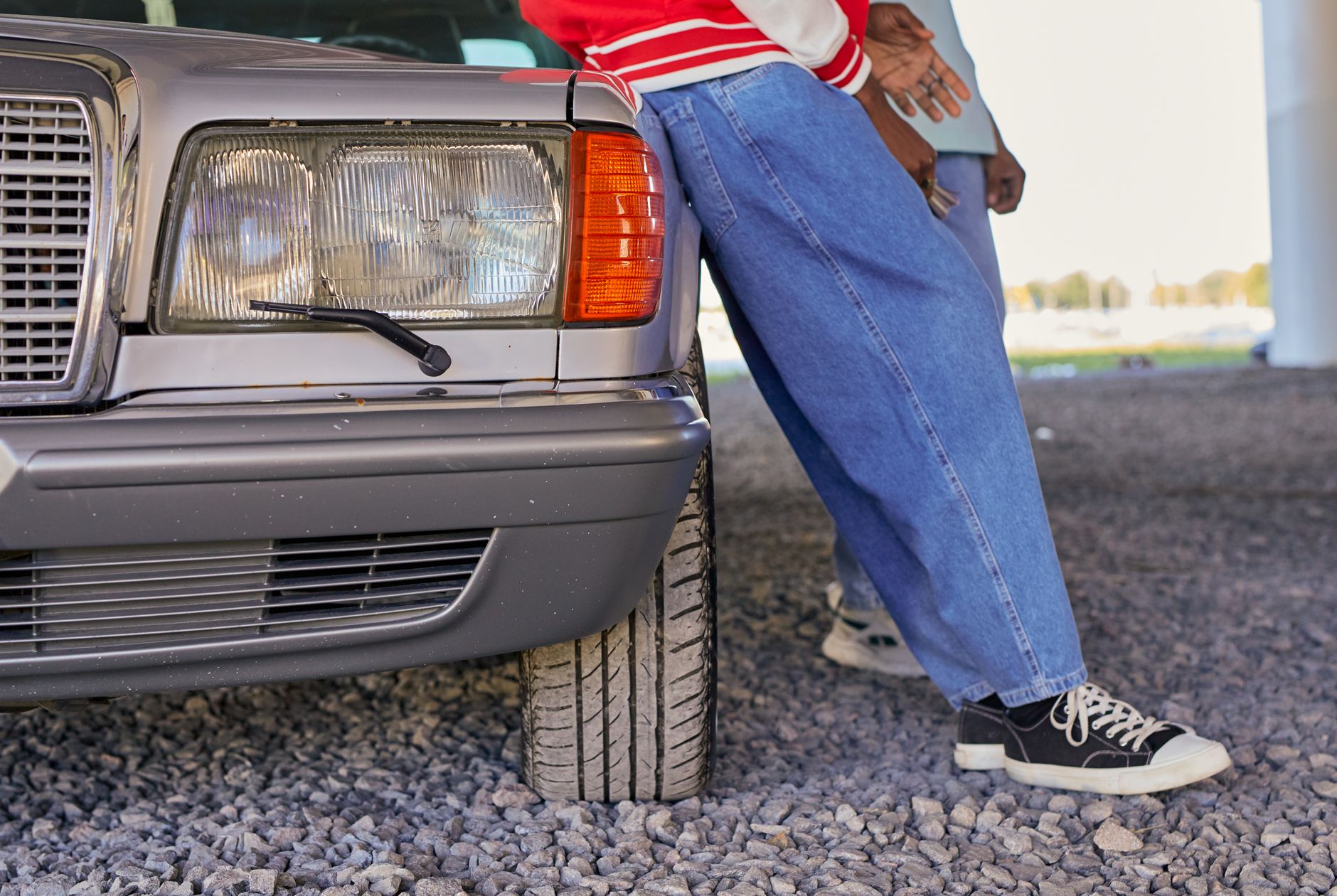 Two people leaning against a vintage Mercedes parked on gravel, showcasing urban style with denim and sneakers.