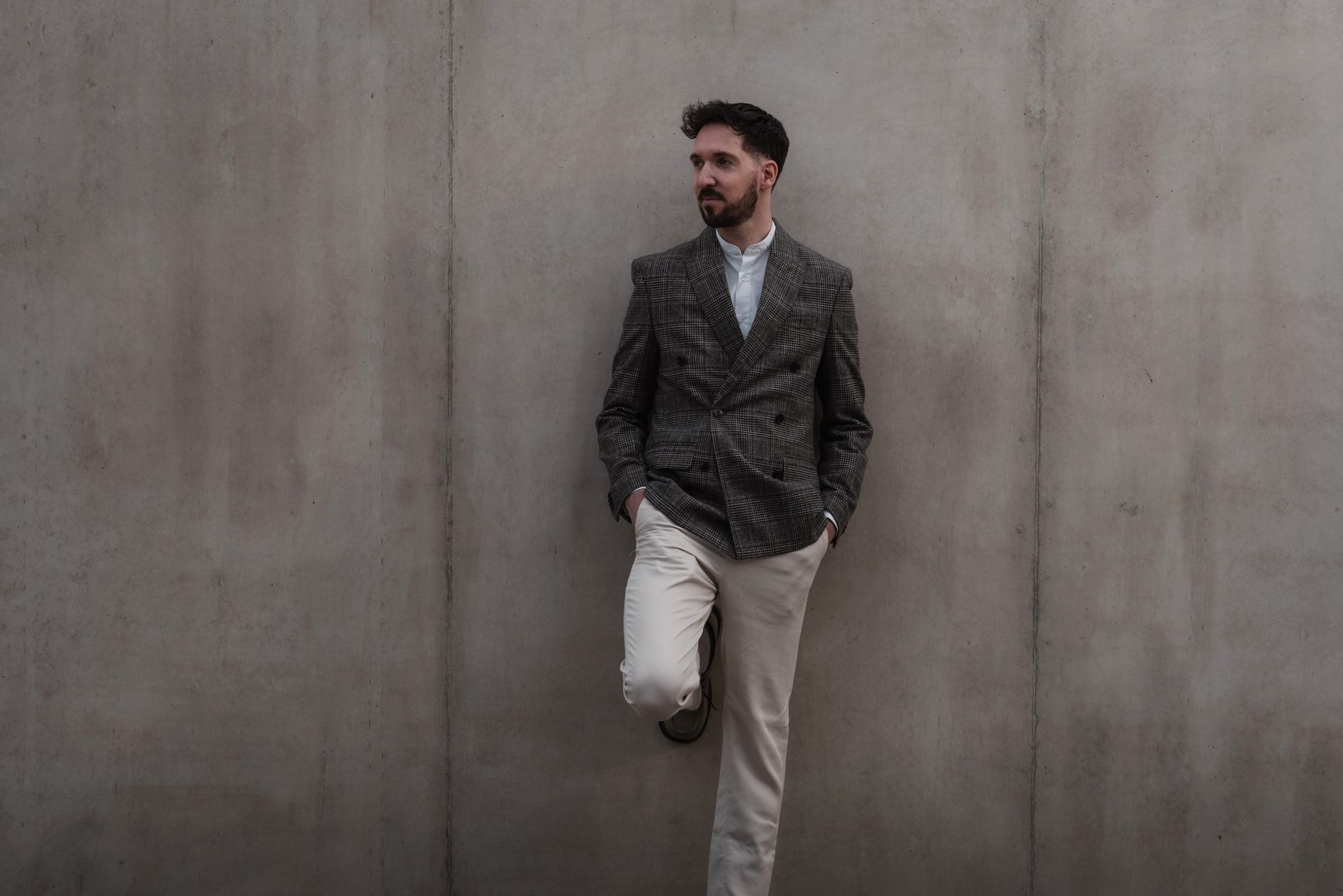 Young man in a checkered blazer leaning against a concrete wall, exuding modern fashion and casual elegance.