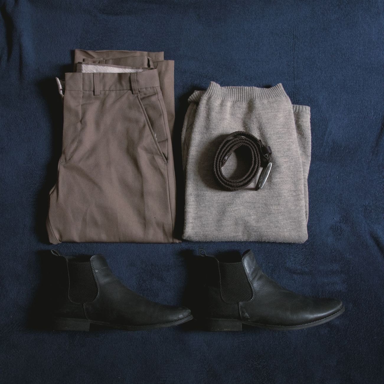 Men's clothing ensemble including trousers, sweater, boots, and belt arranged on a blue surface.