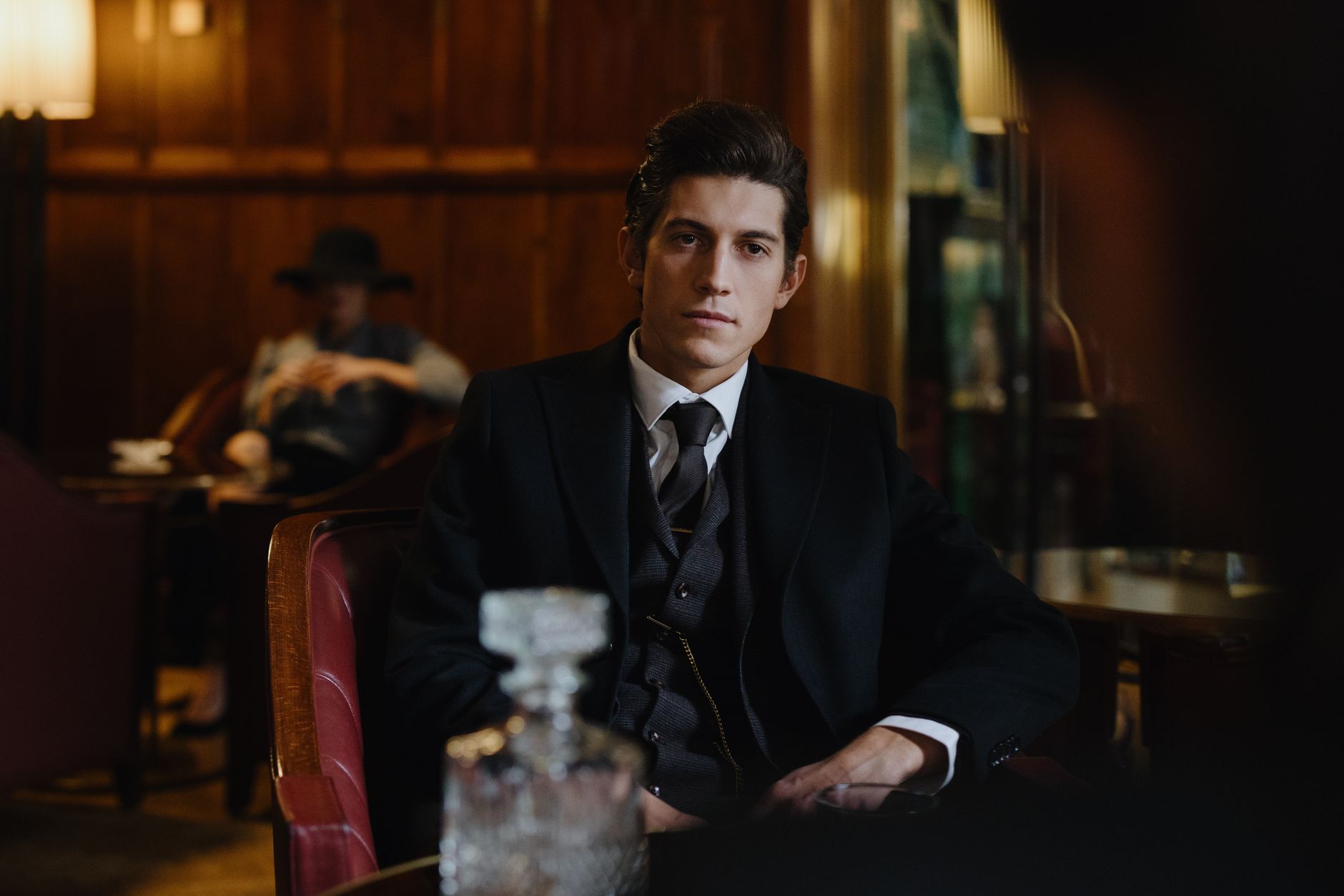 A stylish man in a classic suit sits elegantly in a vintage restaurant setting, exuding sophistication.