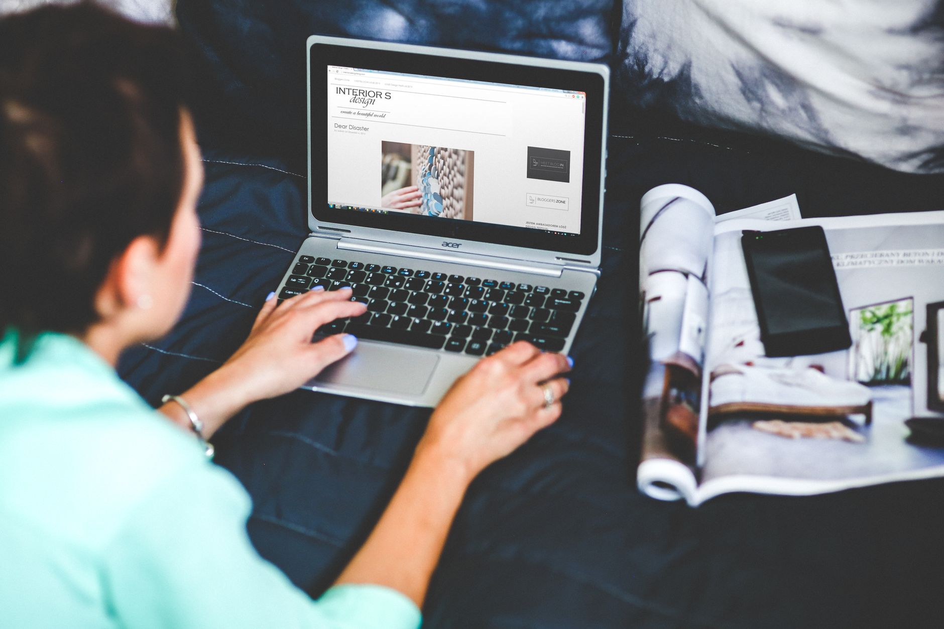 A woman on a bed browsing and blogging with a laptop and a magazine nearby.