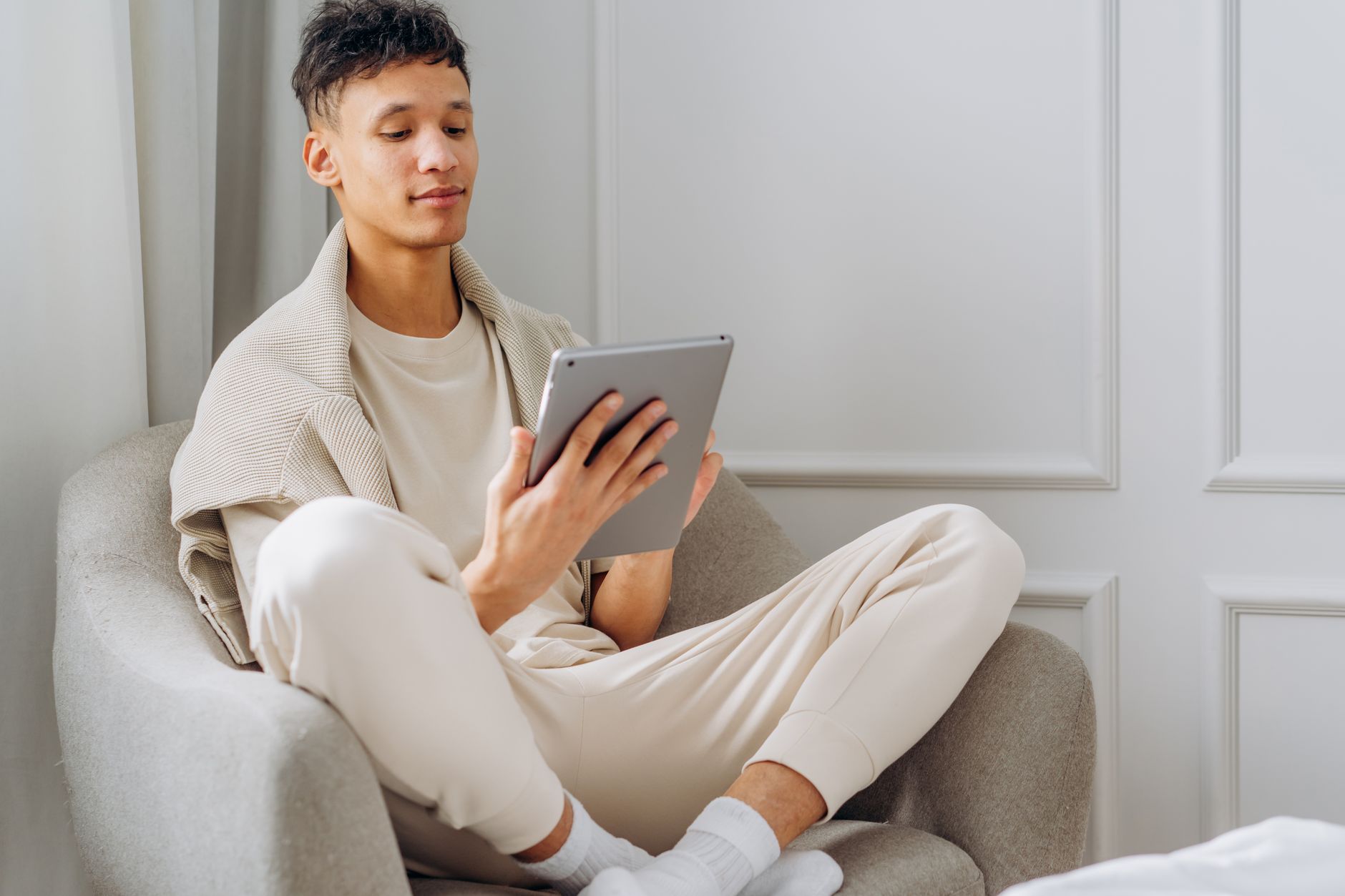 A young man comfortably sits on a chair indoors, reading on his tablet.