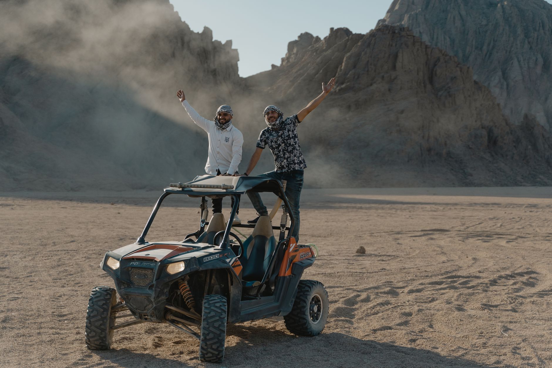 Two men riding a dune buggy in a rocky desert landscape with hills in the background, celebrating their adventure.