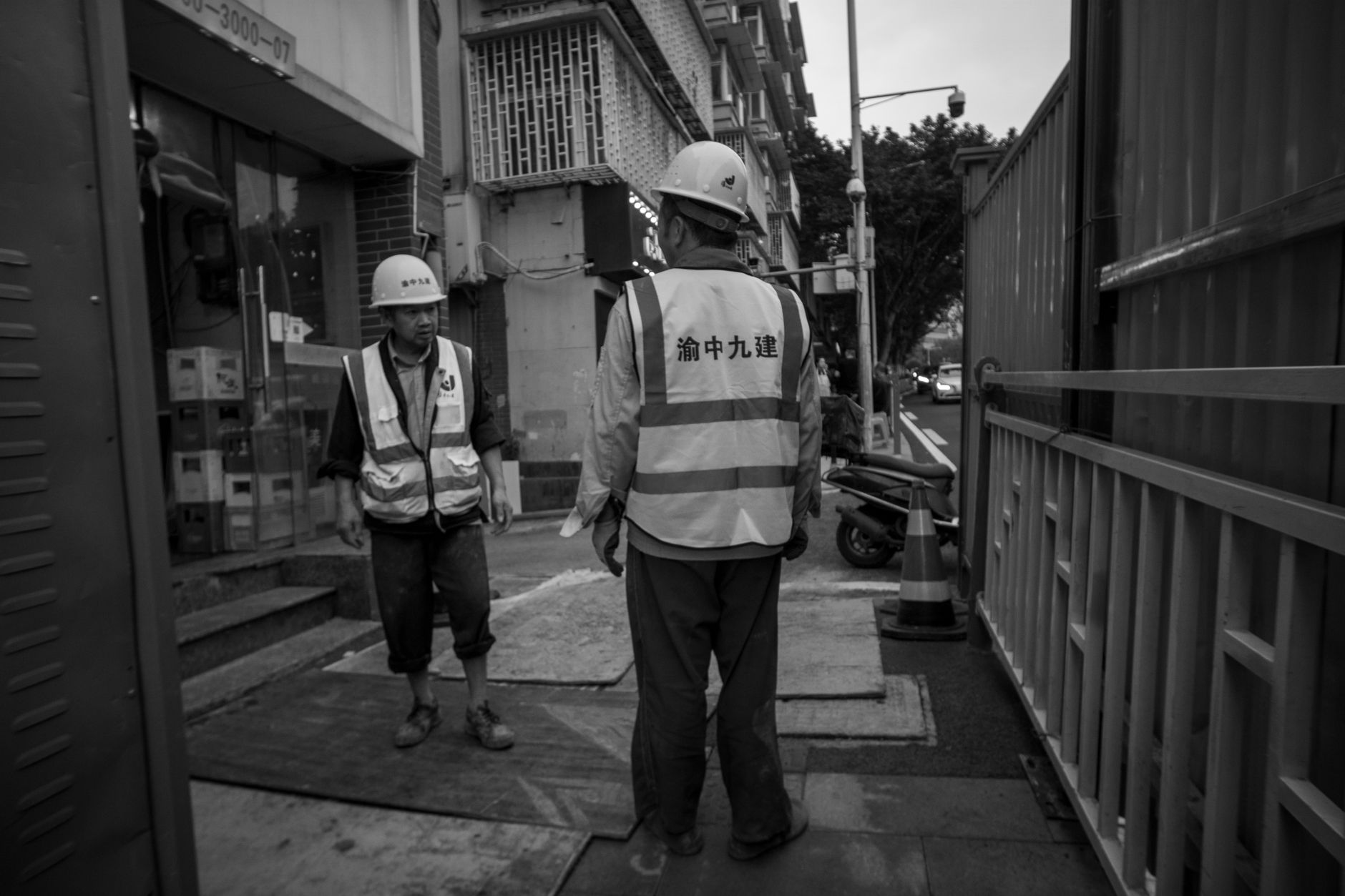 Two construction workers in reflective vests and helmets stand on an urban street.
