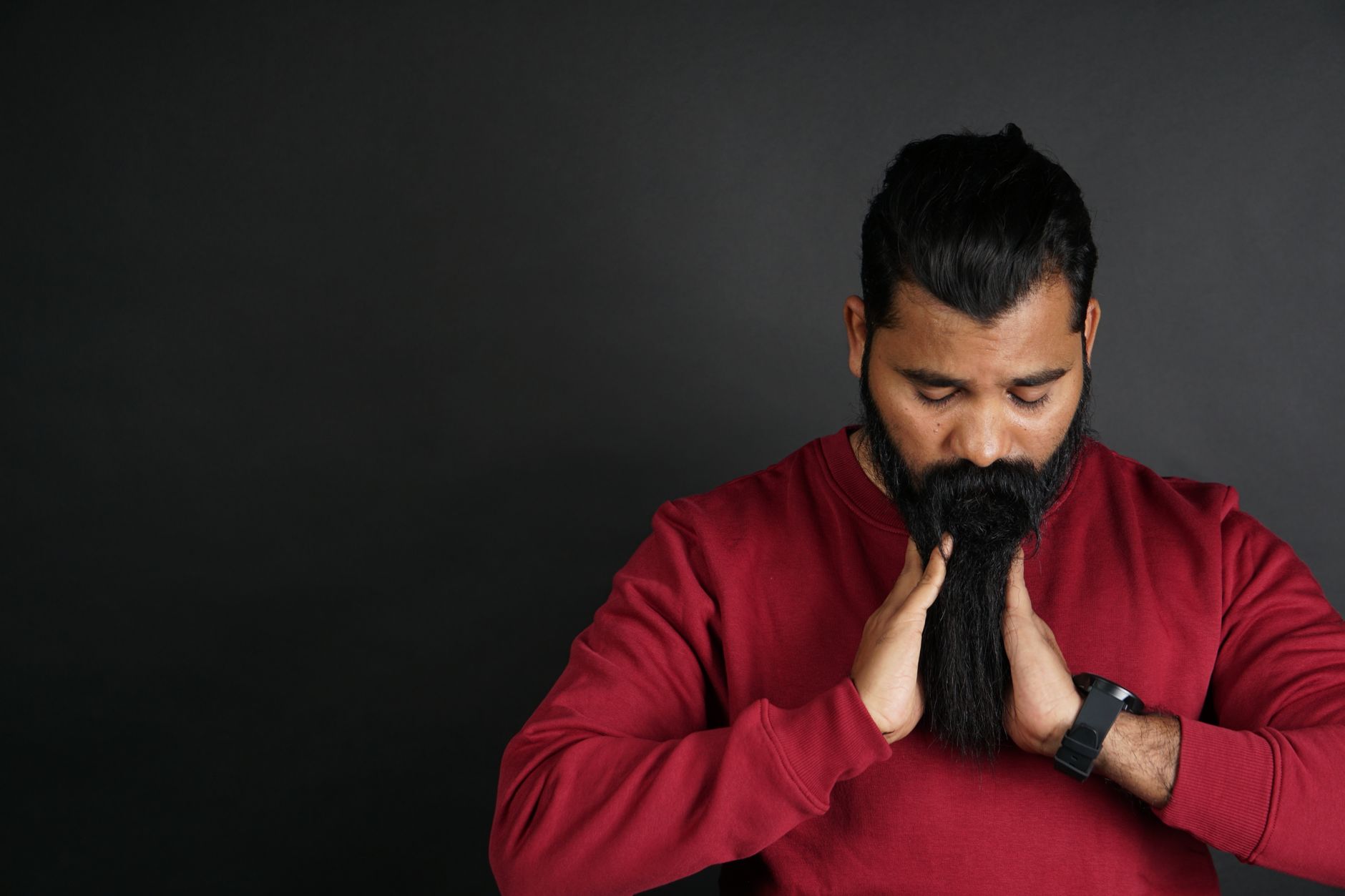 Portrait of a man with a long beard in a red sweater adjusting facial hair in a studio setting.