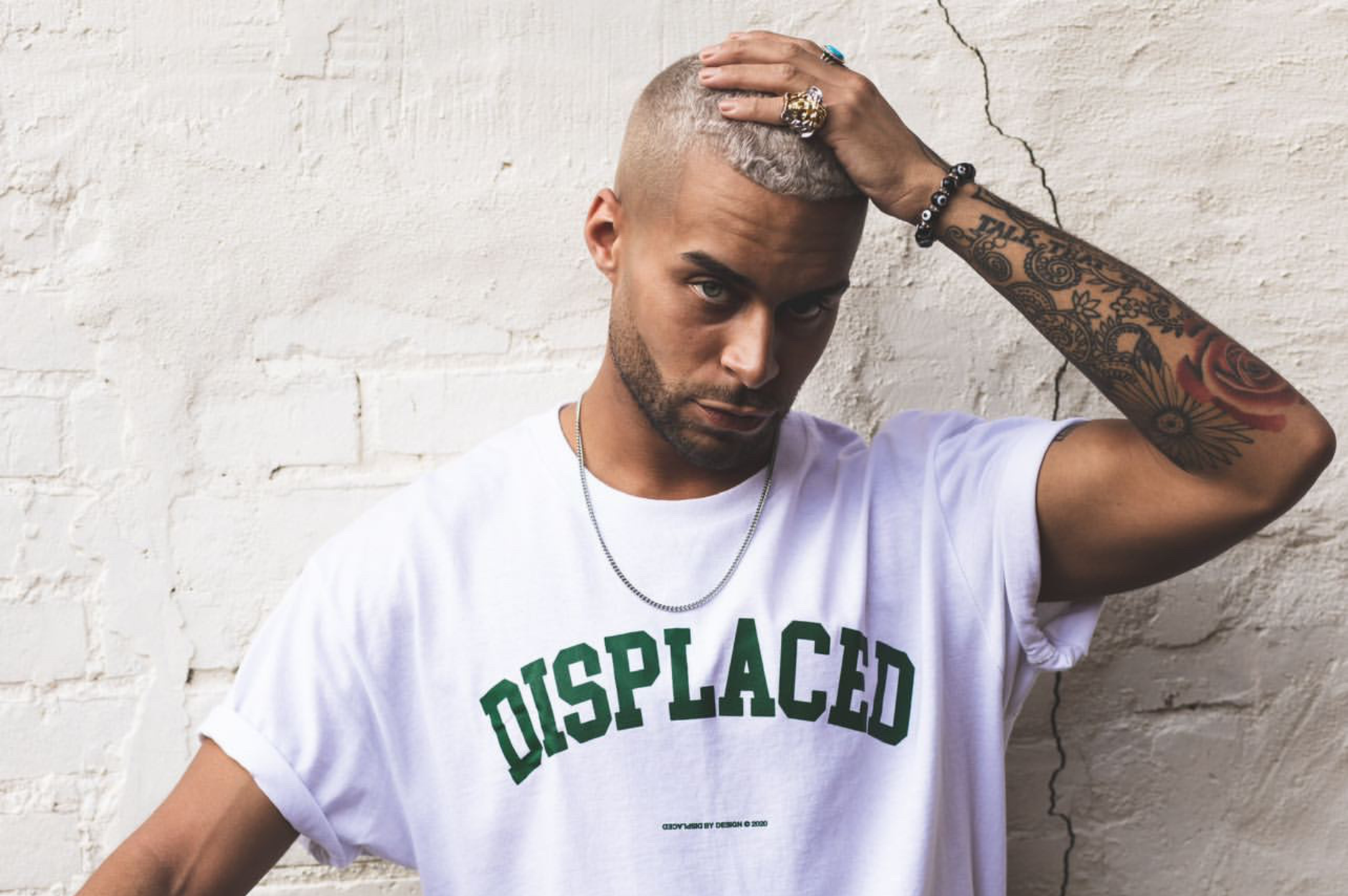 Stylish man wearing 'Displaced' t-shirt and jewelry poses confidently against a white brick wall.