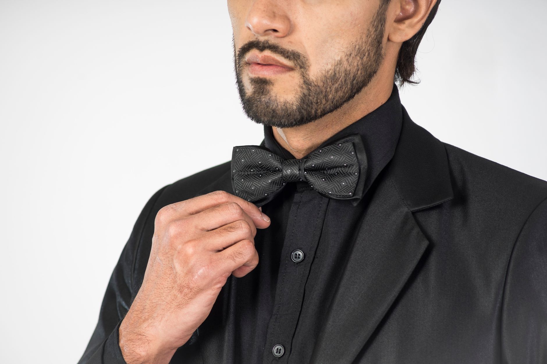 A stylish man in formal black attire adjusts his bow tie against a white background.