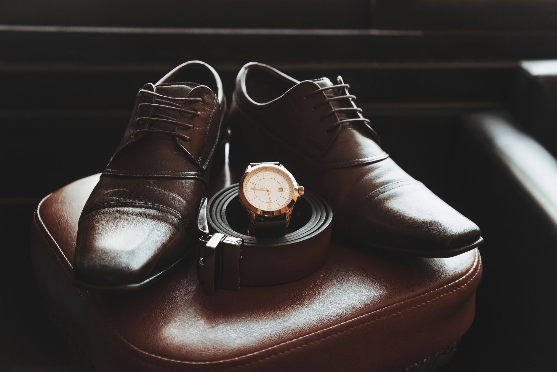Close-up of stylish men's shoes, belt, and wristwatch on a leather chair, showcasing elegance and fashion.