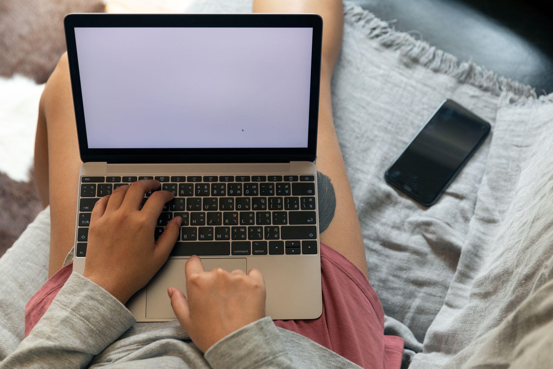 From above of crop faceless female remote employee typing on netbook with empty screen while sitting near smartphone in apartment