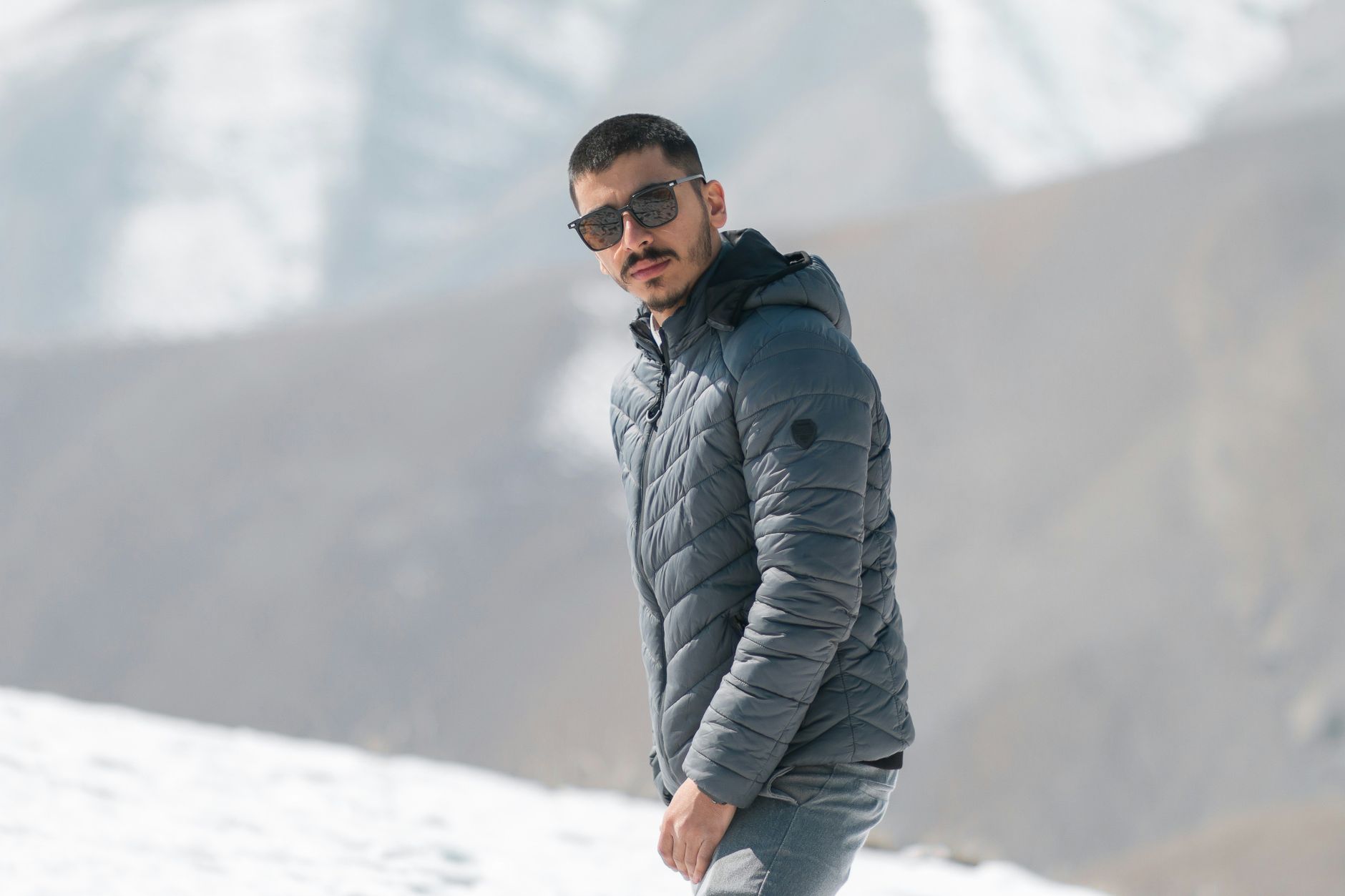 Portrait of a man wearing a winter jacket and sunglasses standing in a snowy mountain setting.